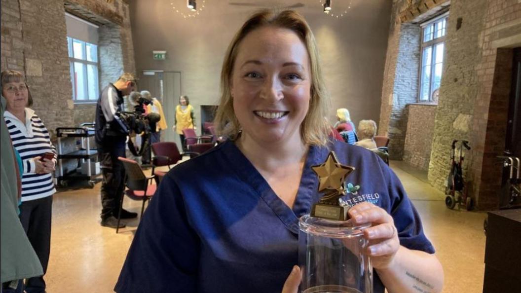 An image of a woman with short blonde hair, smiling at the camera and holding a small star shaped award. She is wearing a navy blue nurses uniform. In the background can be seen a number of people sitting at tables or standing and watching.