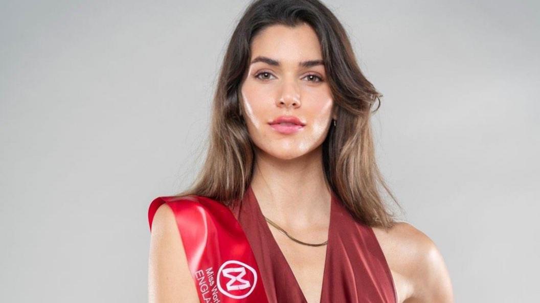 Phoebe looks into the camera as part of a professional photo shoot. She has brown, shoulder length hair and is wearing a red top with a red sash across her shoulder. There is a grey background behind her.