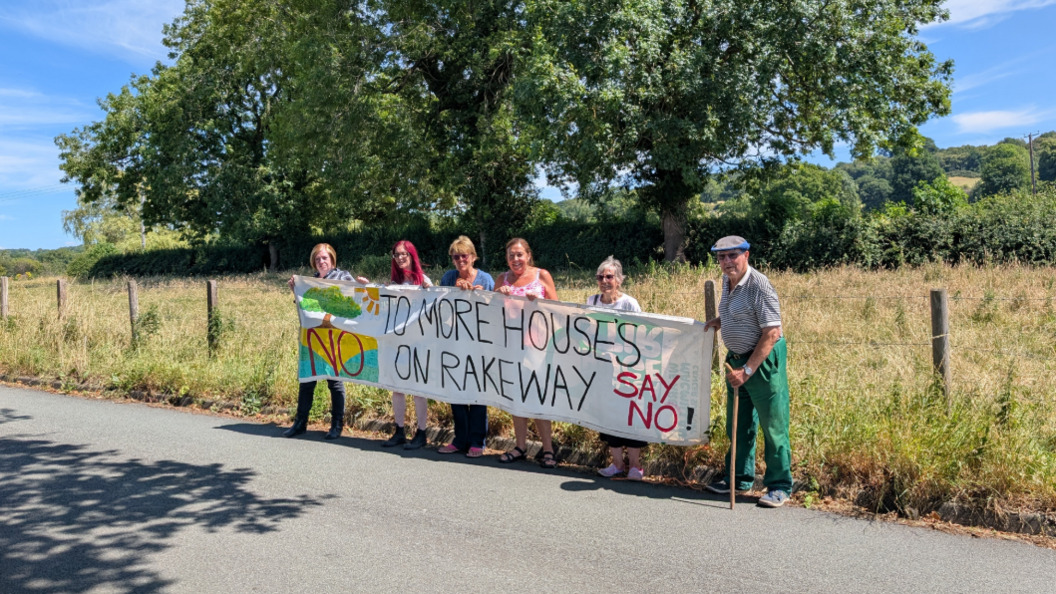 Six people standing on the edge of a road are holding a large banner that states: "No to more houses on Rakeway. Say no!" There is a field and large trees behind them.