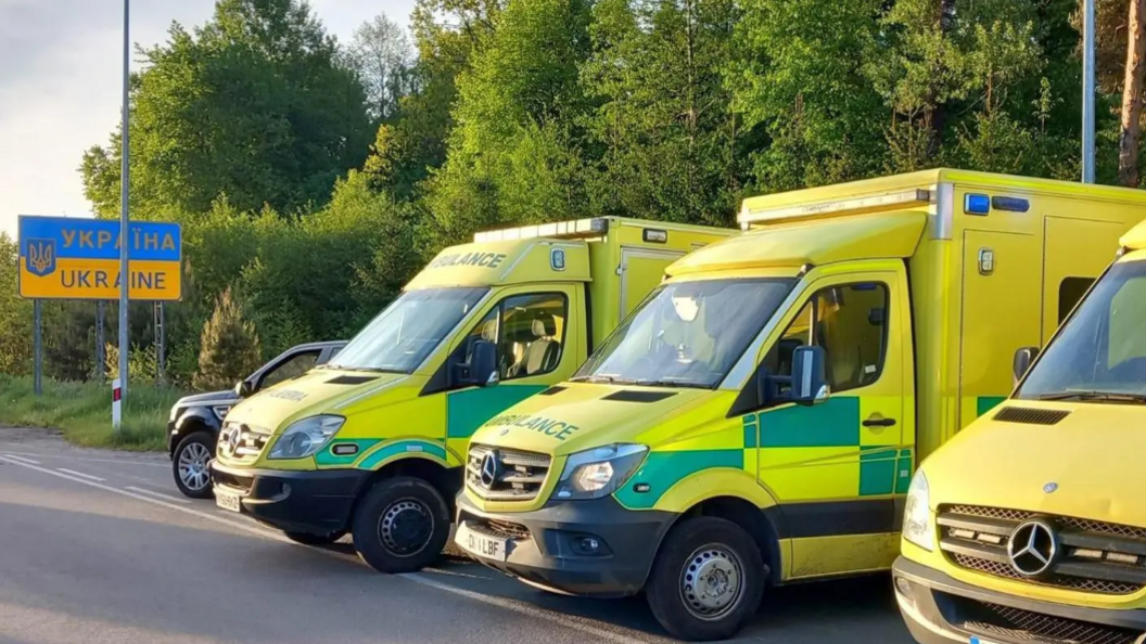 Three ambulances parked on the side of a road, next to a sign for Ukraine. The ambulances are mainly yellow-green with darker green rectangles in places on the side and the word "ambulance" stencilled in green on their bonnet.