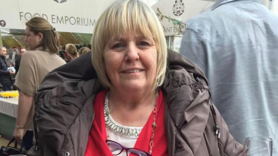 Shelagh Goodwin in a room, with short blond hair, wearing a brown coat, with a top underneath, a red jacket and glasses round her neck, on a chain. There are a number of people behind her, she is smiling and looking straight at the camera.