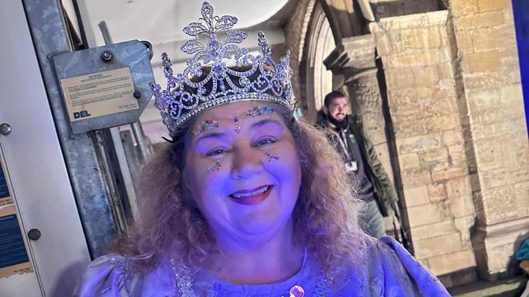 Cheryl Fergison dressed as a fairy godmother, complete with crown, sparkly makeup and dress, standing outside a building in Peterborough, a man can be seen smiling in the background to the right. 