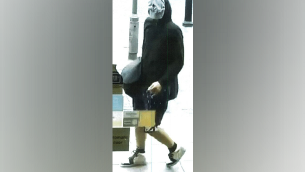A man dressed in a black hoodie and shorts, and trainers, strolls in a shop holding a bag. His hood is up and his face is covered with a white pair of boxer style briefs.