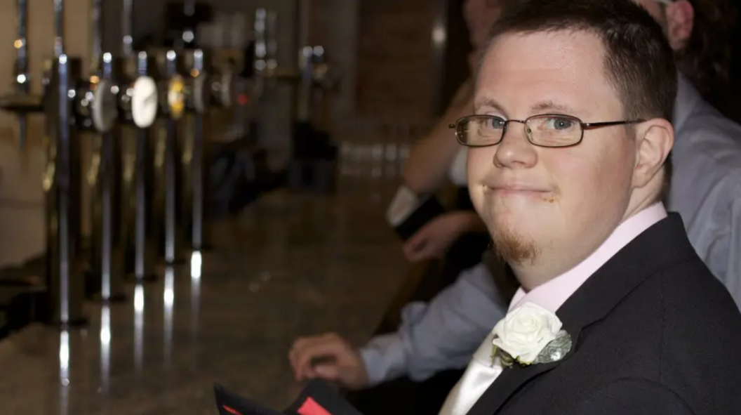 Richard Handley smiles at the camera as he is photographed wearing glasses and a smart suit and tie. He has a white rose attached to his lapel. He is standing a bar. 