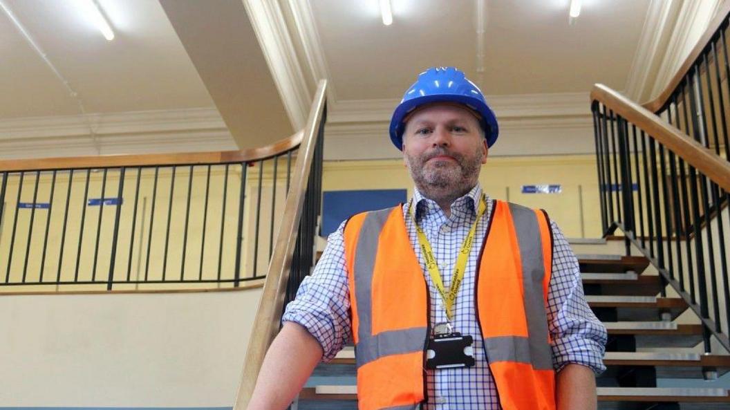 Museums head Damian Etheraads at the site of the new Hereford museum and gallery, where building work is due to begin shortly. He is standing on a staircase and has a hi-vis jacket, a yellow lanyard and a helmet.