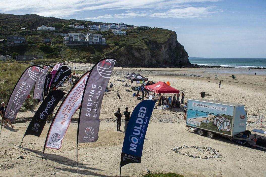 A sandy beach on a sunny day, surrounded by cliffs. Surfing banners and a Surfing England hut are high up the beach, along with gazebos featuring surfers and equipment under them.