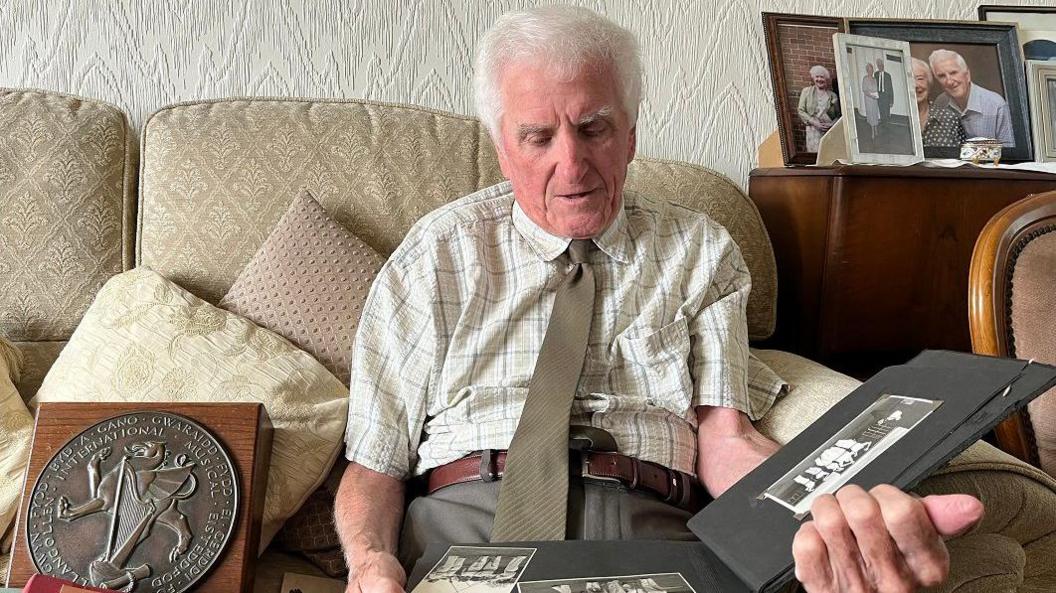 John Hill looking through old pictures, wearing a cotton patterned shirt and brown tie, sitting on a cream sofa. There are pictures of him and his wife in the background and a round plaque trophy next to him.