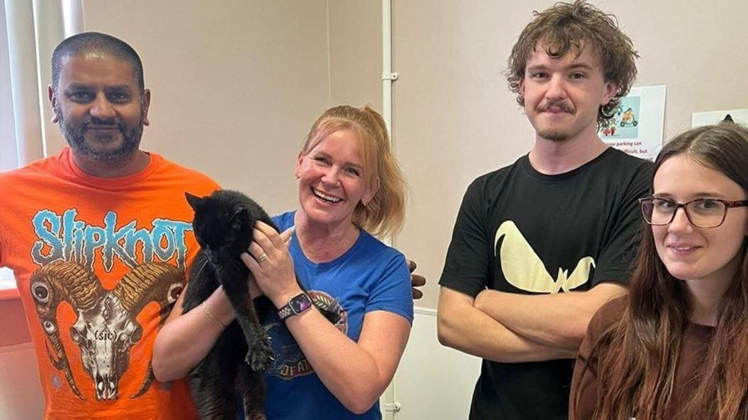 Two men and two women in a room with a black cat. The first man has dark hair and a dark beard and an orange t-shirt, the first woman has blonde hair and a blue t-shirt and is holding the cat. The second man has wavy brown hair and a beard and black t-shirt and the second woman has long brown hair and glasses