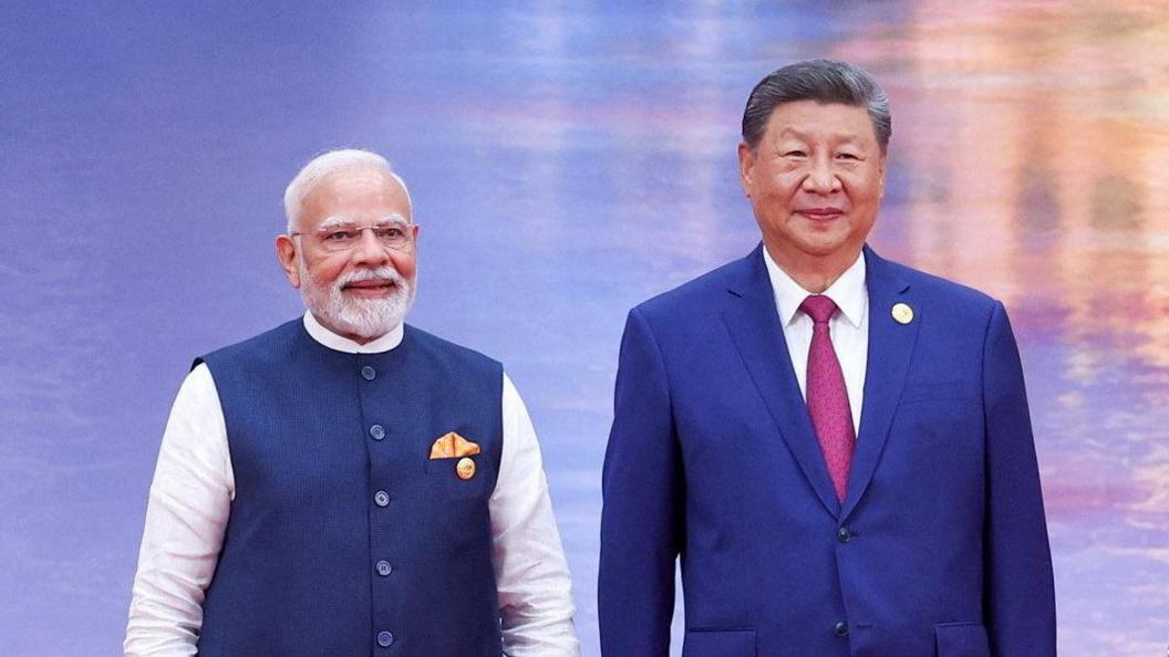 Narendra Modi and Xi Jinping smile for the cameras on stage against a shimmering blue and orange backdrop. Modi is wearing a blue vest over a white kurta with a golden pocket handkerchief, Xi is wearing a navy blue suit and a maroon tie.