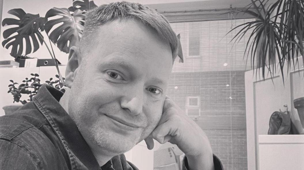 Chris with his head resting on his hand smiling at the camera with a plant in the background