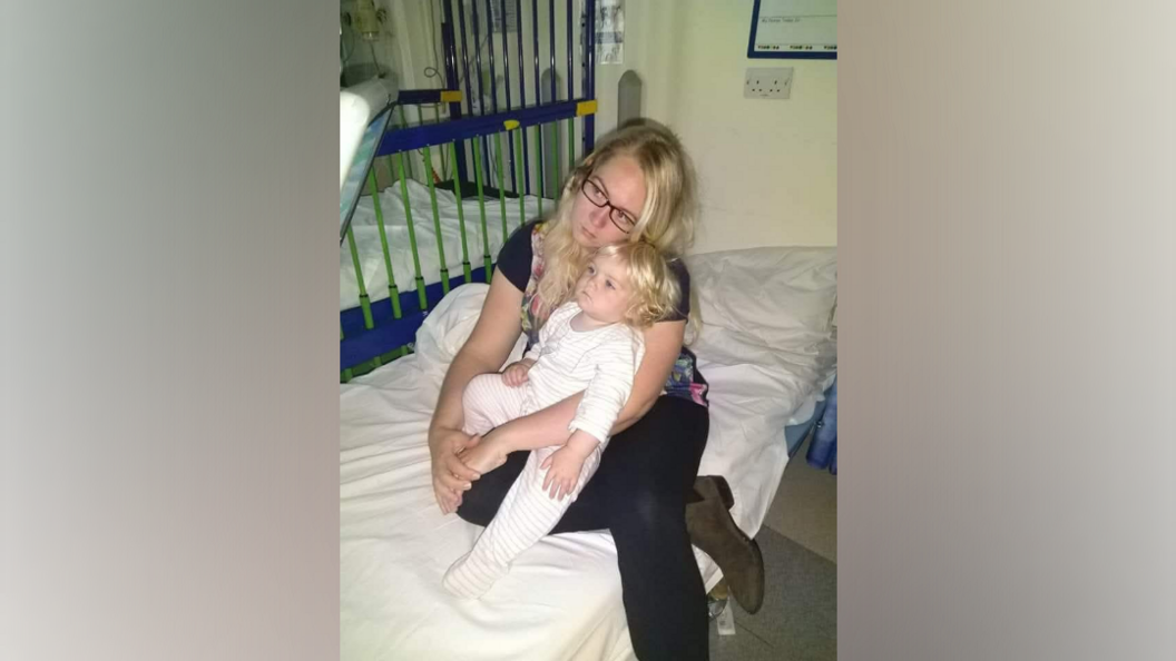 A woman with blonde hair and glasses, looks worried as she cradles a young toddler, with blonde hair, wearing a white baby grow. Both are sitting on a hospital bed