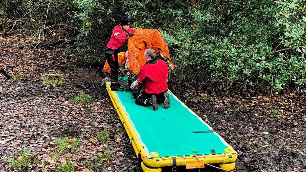 Rescuers in the process of freeing a woman from thick mud deep in the woods, with an inflatable walkway for her behind them