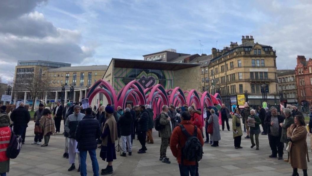 Large gathering of people standing in a group in a pedestrianised square. Behind them is a large pink sculpture and tall sand-coloured buildings in the background.