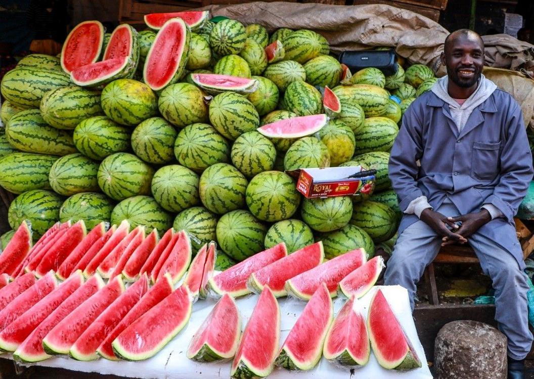 A man sits next to piles of dozens of large melons on his stall.