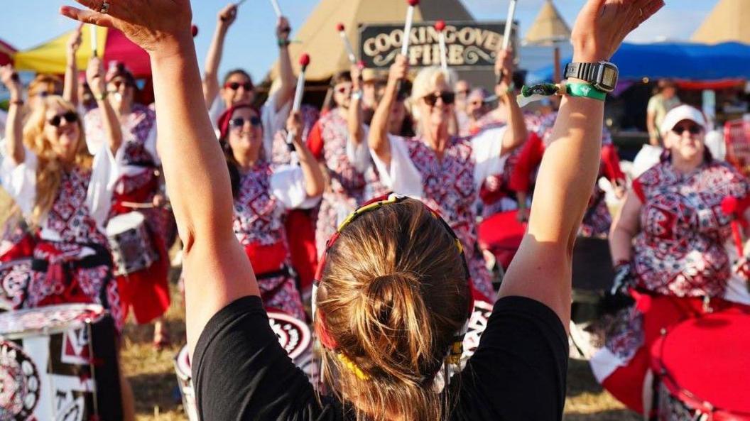 A woman in a black T-shirt holds her hands aloft as she leads a group of drummers at the Lake Paradiso event near Chew Valley Lake. She is facing away from the camera and the drummers, who are all in black and red outfits, are facing towards it