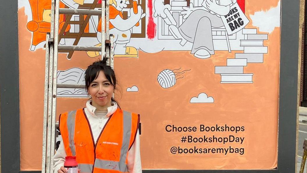 A person in an orange safety vest and paint-splattered jeans stands in front of a large, colourful mural promoting bookshops. The mural, painted in vibrant orange and white, depicts people reading books and includes the text: 'Choose Bookshops #BookshopDay @booksaremybag.' A ladder leans against the mural. The scene is set outdoors on a city street with buildings in the background.