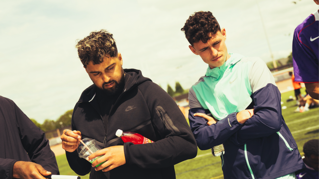 Faraaz Ghaffur and Phil Foden stand by each other at Rico Lewis' charity football tournament, looking down at whiteboards out of the frame. Ghaffur carries two drinks and Foden has his arms folded.