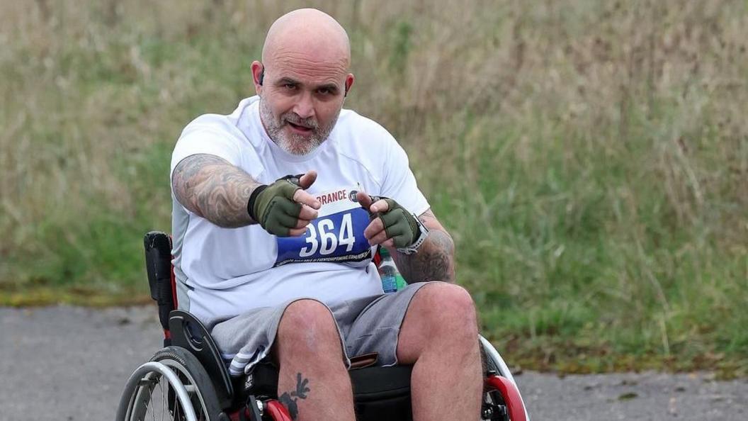 A man in a wheelchair, pointing at the camera and smiling