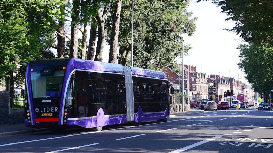 A purple single decker Glider bus on a road lined with trees, ahead on the road are a number of red brick houses and cars stuck at traffic lights. 