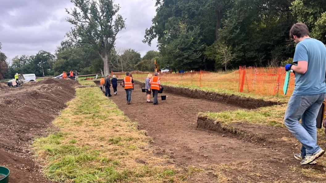 A group of people wearing high vis jackets are standing in a trench with buckets. It is bordered by grass and temporary plastic orange fencing. There are trees in the background.