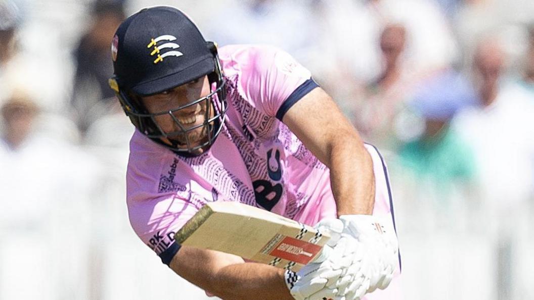 Josh de Caires batting for Middlesex holding his bat out in front of him wearing a pink shirt and helmet