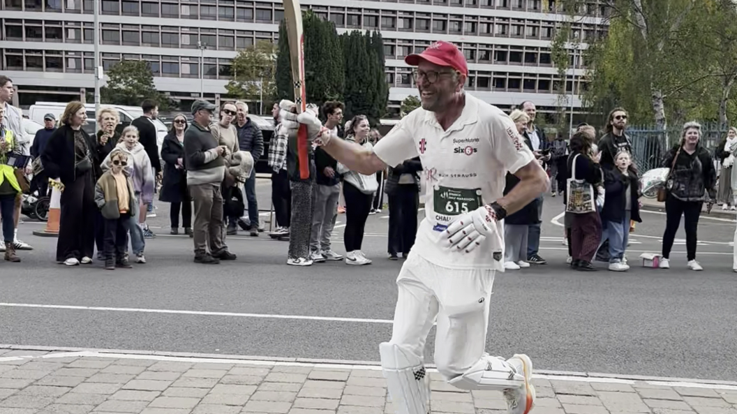 A man wearing a white outfit and holding a cricket bat is running along a road as people cheer