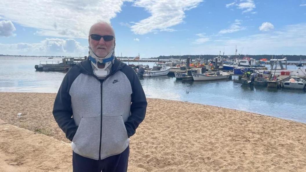 A view of Mr Bascombe in a neck brace, stood on the beach with boats in the background