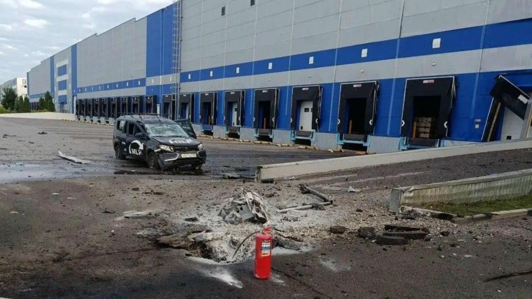 A large building that looks like a warehouse, with multiple entry ports for transport vehicles. The tarmac in front is charred and damaged, and a fire extinguisher has been left at the scene. A car is badly damaged, and the building also appears to have some damage.