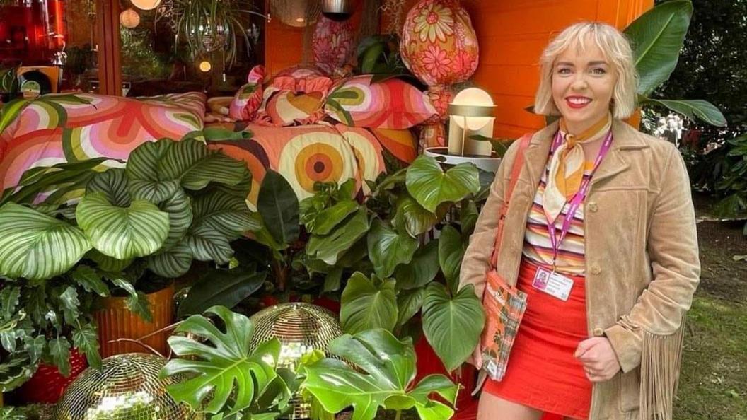 Johanna Haughey-Lewis smiling at the camera.  She hair bobbed blonde hair and is wearing red lipstick, a beige jacket with fringed sleeves, a stripped top, a red mini skirt and a yellow and white scarf.