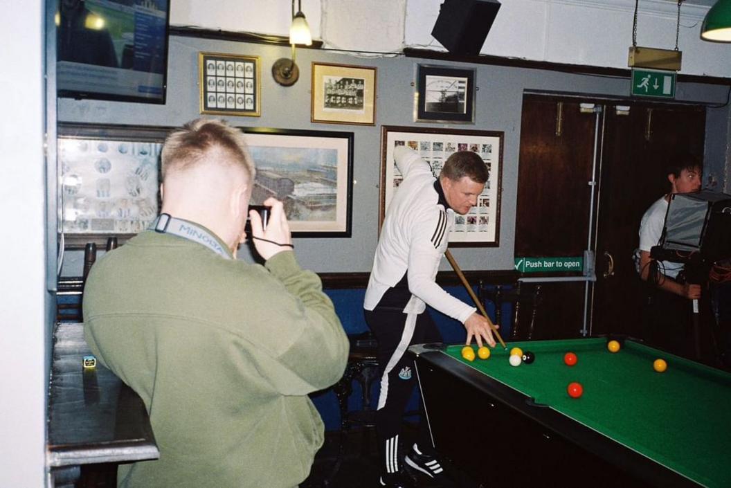 Eddie Howe, who has dark blond hair, is playing pool in a white kit, in the foreground, George Dixon is taking his photo.