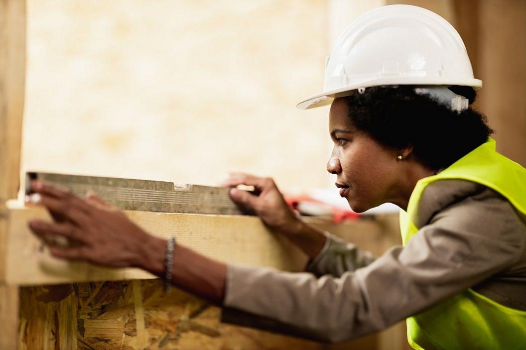 A woman in a hard hat wears a green high-vis jacket over a grey top while looking at a slide rule, a metal bar, on top of a wooden lintel in a window. 