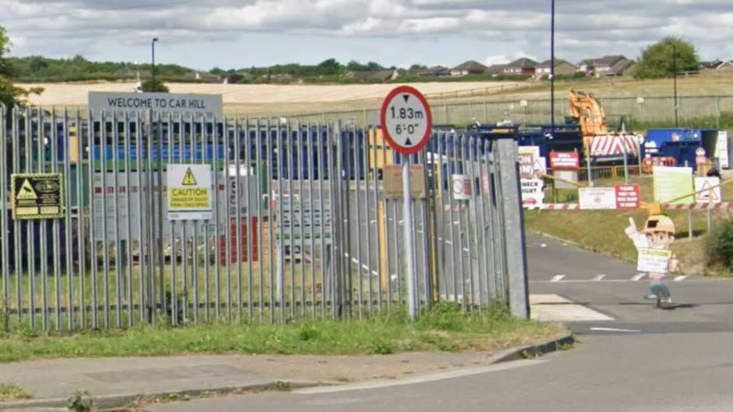 The entrance to a recycling centre. The site has a tall metal fence and there is a red and white barrier across the road entrance.