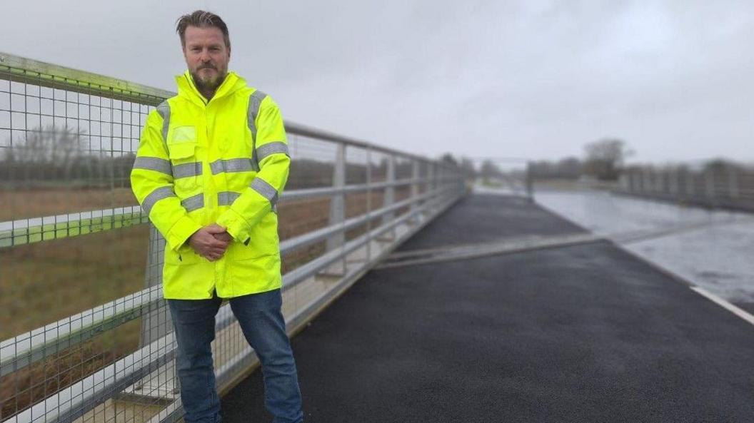 Councillor Chris Watts stood on a bridge on a wet day. He is wearing a yellow his viz jacket, stood next to a barrier. The man has a beard and brown hair. He is wearing jeans.