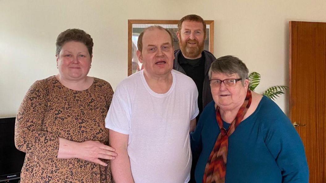 A family group photo of two men and two women standing together in a home.