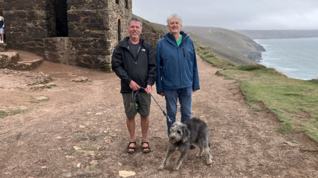 Pat and Richard stood together with Richard's dog Olive. They are stood on the coastal path. The dog is grey and black.