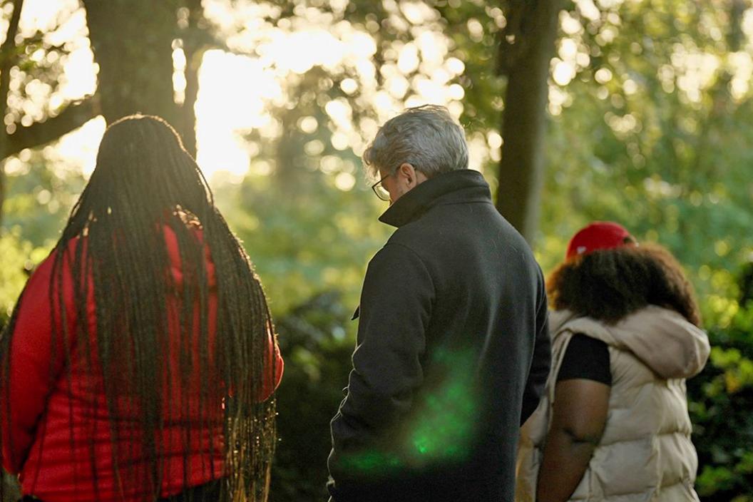 Fergal walks between two South African asylum seekers. Seen from behind, the woman on the left wears a red coat, she has long braids - the woman on the right wears a white sleeveless puffa jacket and red baseball cap over long loose hair