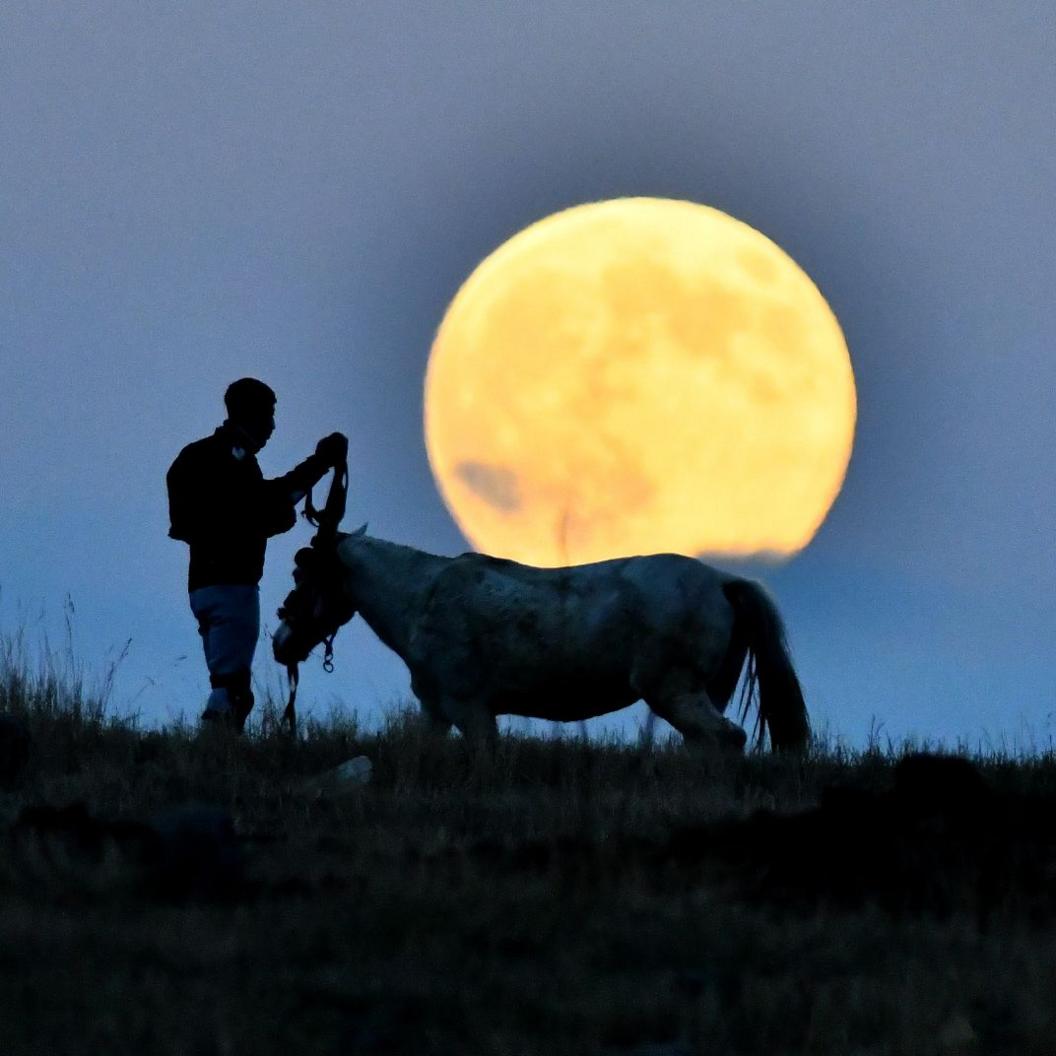 A man and horse silhouette shows him holding reins while standing on a grass hill as evening begins to shroud the scene. The Moon shines a bright orange as it rises in Kars, Turkey on Wednesday.