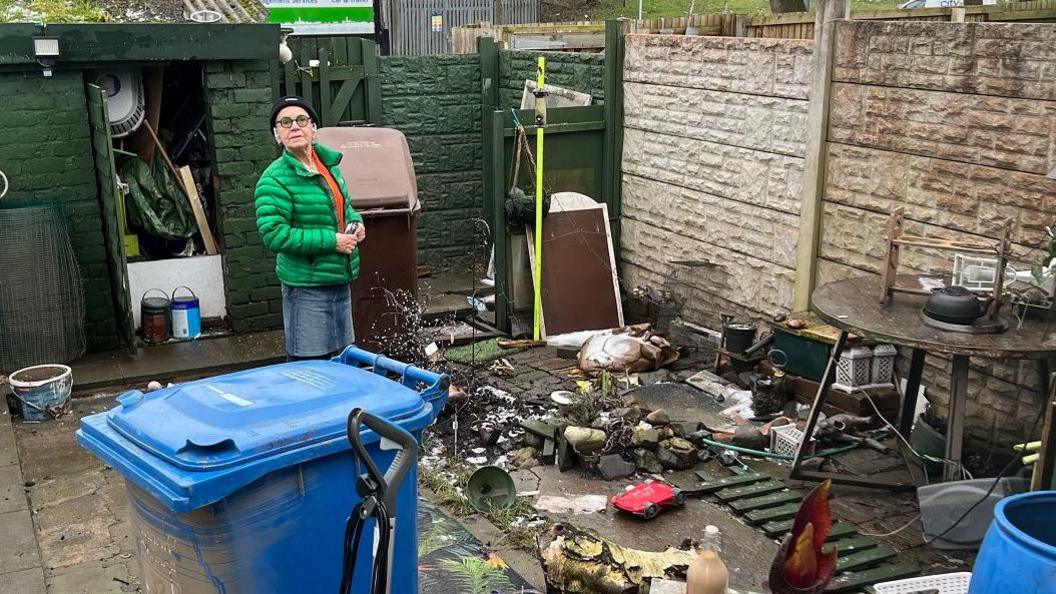 Mia Davy, wearing a green coat, stands in her backyard while clearing up the a pile of flood damaged items outsider her Walthew Lane home