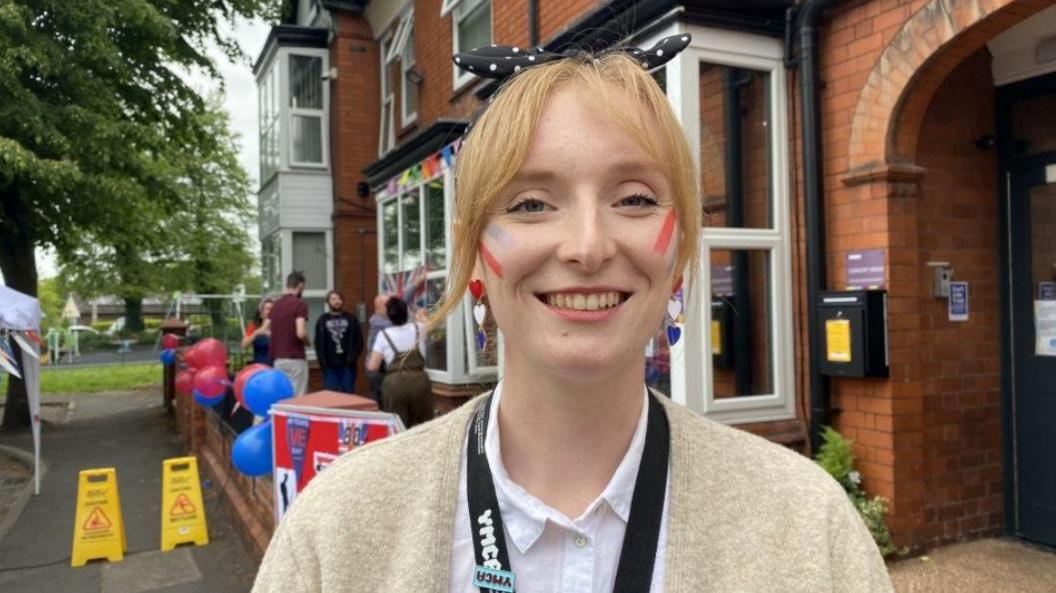 A young woman with tied up ginger hair is wearing a beige cardigan and white shirt and has red and blue stripes on her cheeks. She is stood in a street with red and blue balloons behind her, and there are some other people in the distance.