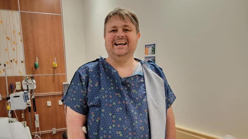 A man stands in a hospital gown and smiles among medical equipment