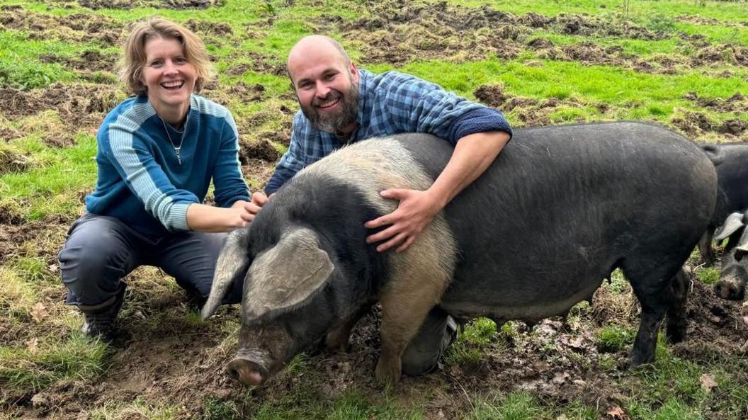 Kate Bigwood with shoulder-length blonde hair and a blue jumper squatting alongside David Gooden, who is bald and has a thick beard. They are next to a large black and white pig called Pepper.