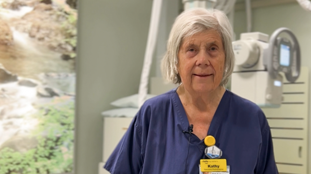 Kathy Thomas wearing blue hospital scrubs and a yellow name tag. She has white hair which is cut just above her jawline, and is smiling at the camera. She is standing in a radiology room at RUH Bath surrounded by medical equipment.