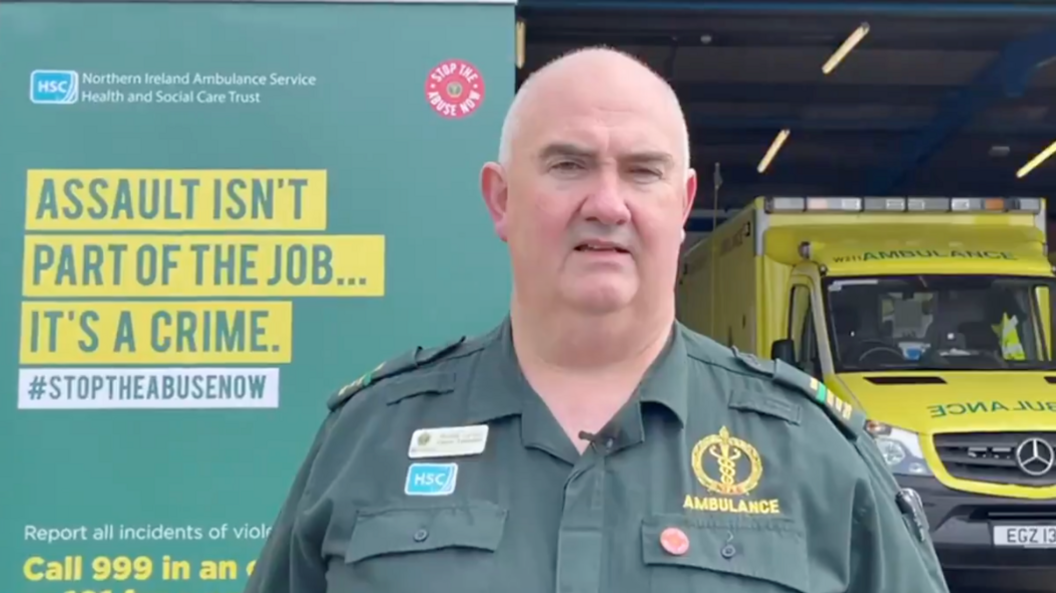 A man in  green ambulance uniform stands in front of a poster against the abuse of emergency staff. Behind him is an ambulance with florescent livery.