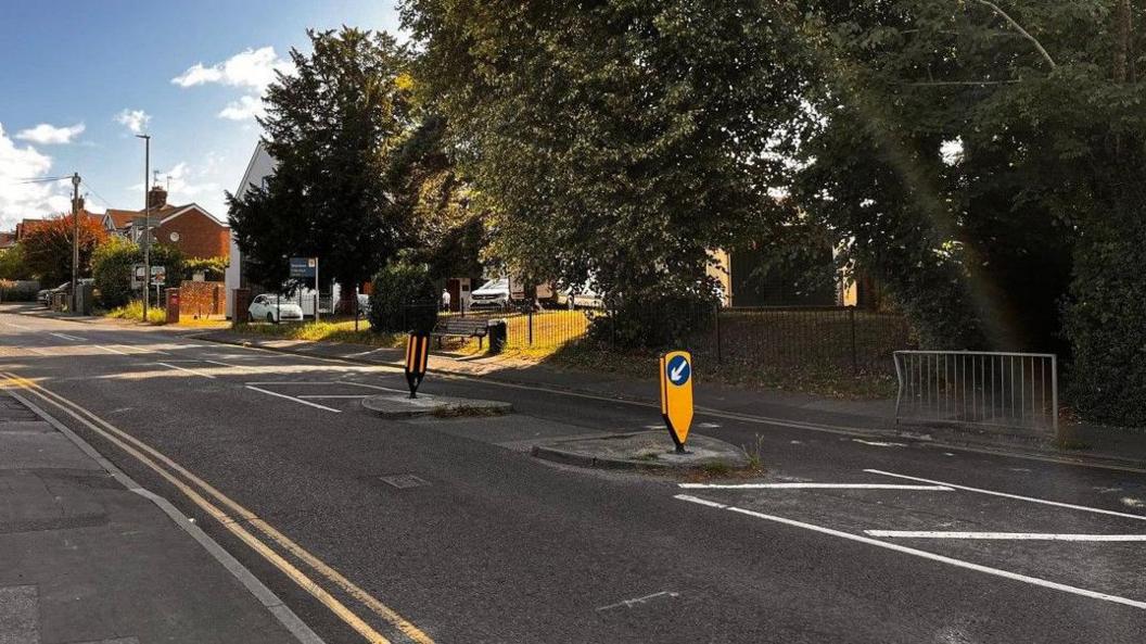 A road with pedestrian island in the centre, double yellow lines down either side of the road and an old metal barrier on the far side of the road