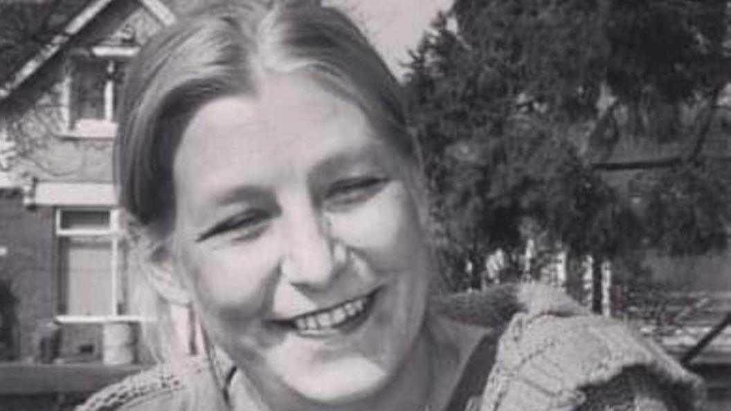 A black and white photo of Dawn Sturgess who is smiling as she sits outside by some trees and buildings