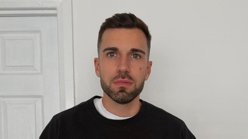 Matthew Connor with short brown hair and a brown beard wearing a black round neck jumper with a white t shirt underneath