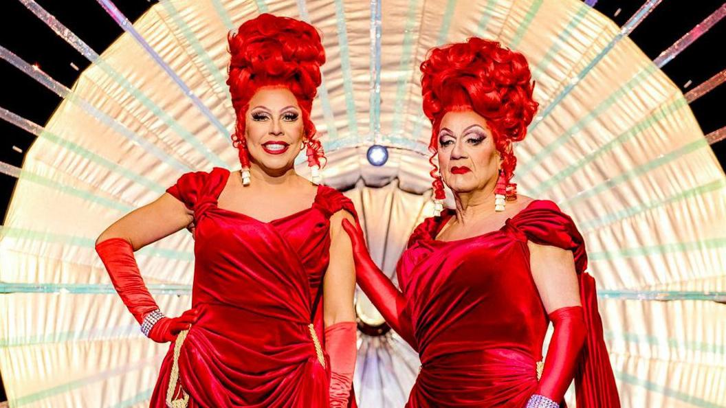 La Voix on stage with her dad Richard Dennis - AKA Voila - in drag. The pair were red wigs piled high, matched with red velvet dresses inspired by theatre curtains and matching red gloves. They're on the runway of RuPaul's Drag Race, with Richard resting his hand on La Voix's arm. 