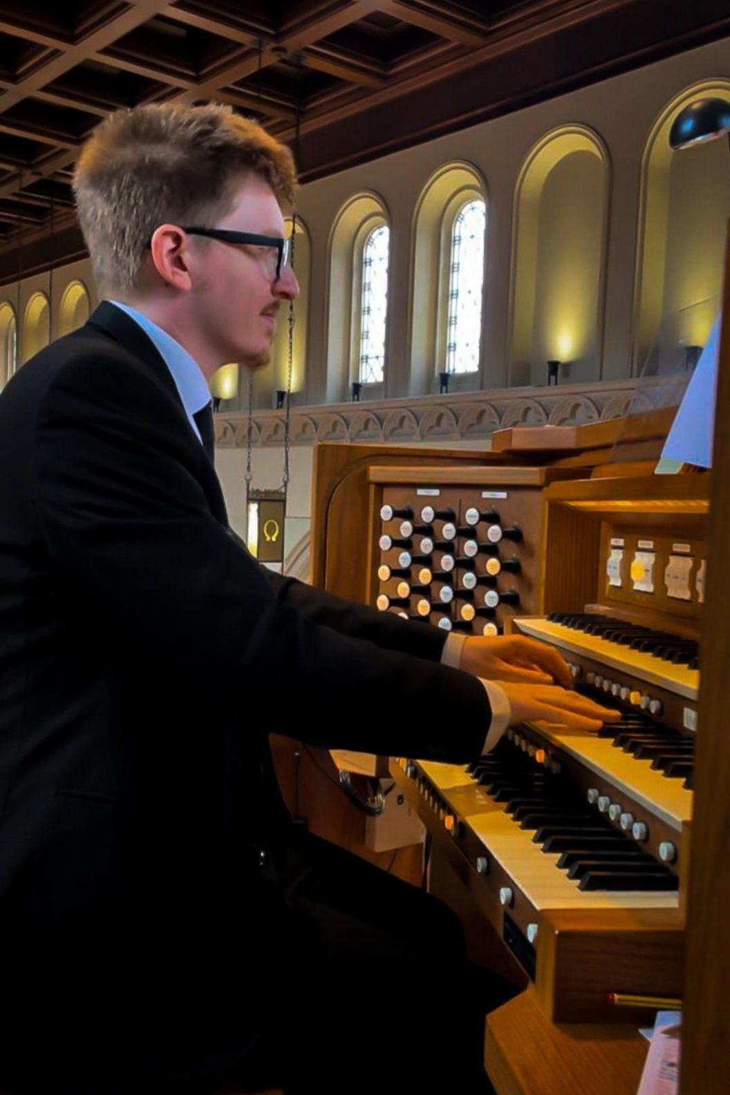 A side-on image of a ginger man in a black suit playing an organ