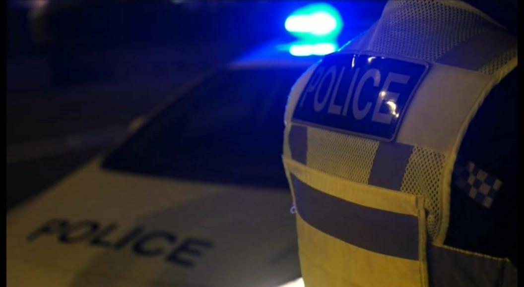 A police officer in a high visibility jacket standing by a police car with its blue light.