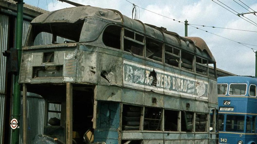 A derelict, grey vintage double-decker bus. Behind it is a restored blue bus.
